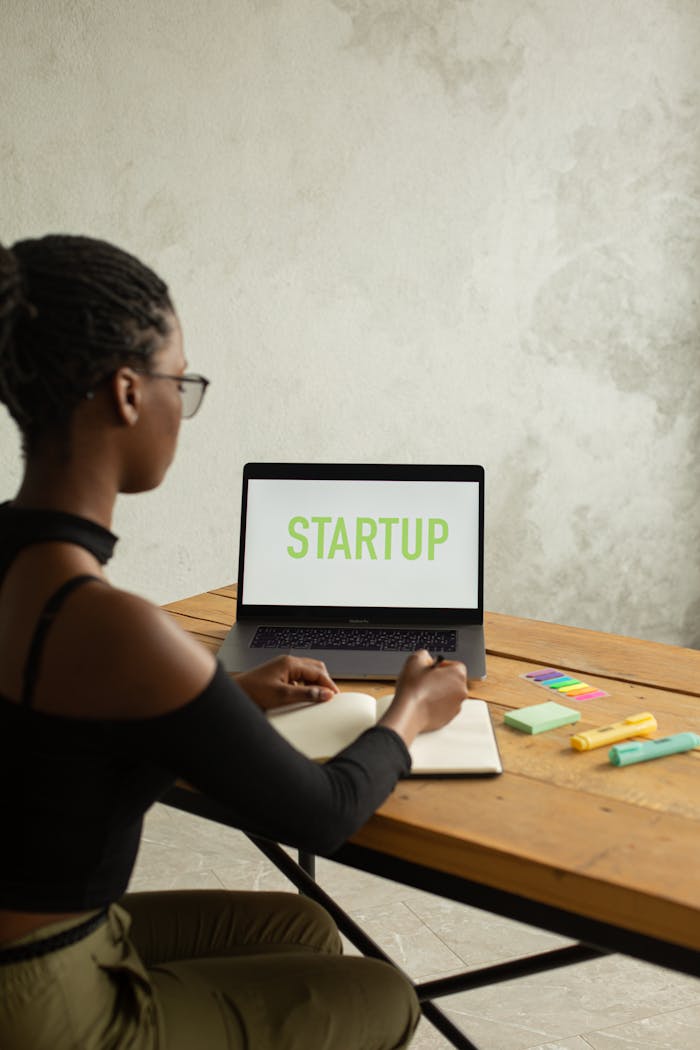 Home Side view of anonymous African American female entrepreneur sitting at table with netbook with startup inscription on screen and writing in notebook using pen and markers